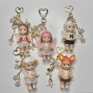 Puede incluir: Una colección de cinco figuras en miniatura, cada una con un disfraz temático de animales único y adjunto a un llavero. Las figuras presentan alas de ángel, faldas y detalles decorativos. Los llaveros tienen cierres en forma de corazón y abalorios.