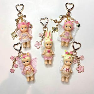 Op de afbeelding: Zes sleutelhangers met kleine, poppenachtige figuren, elk met vleugels en bloemaccenten. Elke sleutelhanger heeft een hartvormige sluiting en gouden en roze bedels. De figuren zijn gekleed in pastelkleuren.