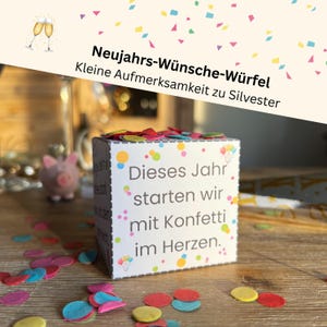 May include: A white cube with colorful confetti and text in German: "Dieses Jahr starten wir mit Konfetti im Herzen." The cube is decorated with colorful dots and is on a wooden surface. The image also includes a small pink piggy bank and two champagne glasses.