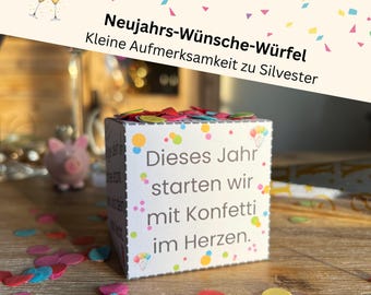 May include: A white cube with colorful confetti and text in German: "Dieses Jahr starten wir mit Konfetti im Herzen." The cube is decorated with colorful dots and is on a wooden surface. The image also includes a small pink piggy bank and two champagne glasses.