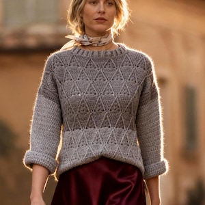 Op de afbeelding: Een vrouw draagt een grijze gebreide trui met een ruitpatroon en een bordeauxrode satijnen midi-rok. Ze draagt bruine leren enkellaarsjes en een sjaal met patroon. De foto is buiten genomen op een geplaveide straat.