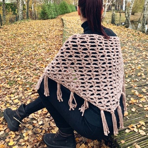 May include: A woman wearing a pink crocheted shawl with fringe. The shawl is a triangular shape and has a delicate openwork pattern. The woman is sitting on a path covered in fallen leaves.