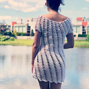 Puede incluir: Un vestido de crochet blanco de corte holgado y mangas cortas. El vestido está hecho de una tela delicada y de tejido abierto.