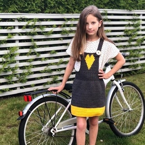 Puede incluir: Una niña con un vestido de crochet negro y amarillo con un diseño en forma de corazón, de pie junto a una bicicleta.