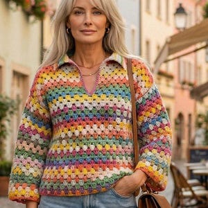 Op de afbeelding: Een veelkleurige gehaakte trui met een V-hals en lange mouwen. De trui heeft regenboogkleuren en wordt gecombineerd met lichtblauwe jeans en een bruine handtas. De vrouw staat op een geplaveide straat.