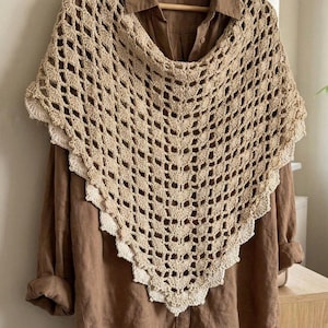 Puede incluir: Un chal de ganchillo beige con un delicado diseño calado, sobre una camisa marrón. El chal tiene un borde festoneado y está colgado de una percha de madera.