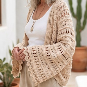 May include: A beige crochet cardigan with openwork detailing and flared sleeves. The cardigan is worn over a white tank top and paired with beige pants. A long necklace with a pendant is also visible.