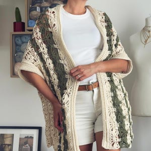 May include: A crochet cardigan featuring an openwork pattern in cream and olive green yarn. The garment has short sleeves, a straight hem, and a solid cream-coloured border along the front opening, worn over a white t-shirt and white shorts.