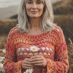 Puede incluir: Un suéter de ganchillo en tonos naranja y rojo, con una flor de margarita. El diseño de tejido abierto revela una capa inferior blanca. El suéter tiene mangas largas y cuello redondo. La mujer de la imagen sostiene una flor de margarita.