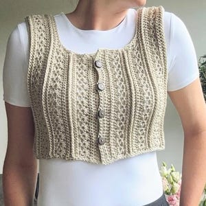Könnte beinhalten: Eine beige, gecroppte Weste mit Knöpfen und quadratischem Ausschnitt. Die Weste hat ein strukturiertes, offen gewebtes Design und wird über einem weißen Oberteil getragen. Die Knöpfe sind dunkel und neutral. Der Gesamtstil ist lässig und handgefertigt.