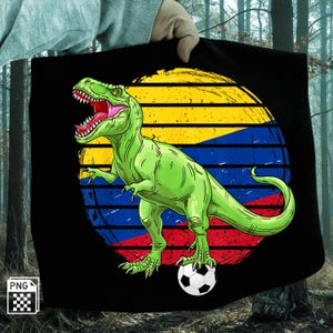 Pode incluir: Camiseta preta com um desenho de um Tiranossauro Rex verde, com a boca aberta, em pé sobre uma bola de futebol. O design inclui um fundo circular com listras amarelas, azuis e vermelhas.
