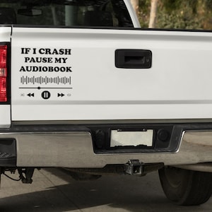 Pode incluir: Porta traseira de caminhão branca com um decalque preto que diz "IF I CRASH PAUSE MY AUDIOBOOK" acima de um gráfico de forma de onda de áudio e ícones de controle de mídia. O caminhão tem lanternas traseiras vermelhas e um para-choque preto.