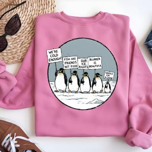 May include: Pink sweatshirt featuring a cartoon of penguins protesting on an icy landscape. The penguins hold signs that read "We're Cold Enough!" and "Fish are Friends, Not Food." A smaller penguin holds a sign that says "Waddle for a Cause."