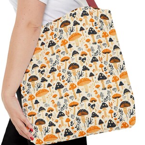 May include: A tote bag featuring a repeating pattern of various mushrooms in shades of orange, brown, and black against a cream background. The bag has a solid dark red shoulder strap and is constructed from a fabric material with a rectangular shape.
