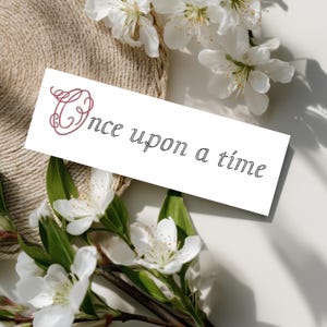Könnte beinhalten: Eine weiße rechteckige Karte mit dem Schriftzug "Once upon a time" in Schreibschrift. Der Buchstabe "O" ist mit einem bordeauxfarbenen Blumendesign verziert. Weiße Blüten und grüne Blätter umgeben die Karte, vor einem beigefarbenen, gewebten Hintergrund.
