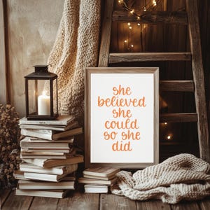 Può includere: Una cornice rettangolare in legno contenente una stampa bianca con il testo arancione 'she believed she could so she did'. La stampa incorniciata è posizionata accanto a una pila di libri, una lanterna in metallo nero con una candela bianca e un tessuto lavorato a maglia.