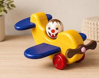 May include: A wooden toy airplane featuring a bright yellow body, blue wings, and a dark brown propeller with a pink center. The cockpit contains a round wooden figure with a painted face, brown hair, blue eyes, a red nose, and two white teeth.