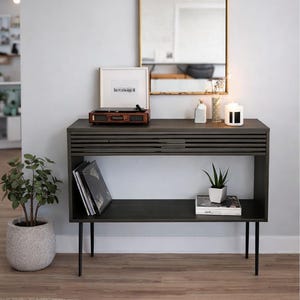 May include: A dark gray console table with a built-in shelf and black legs. A framed picture, a record player, and a candle sit on top. A potted plant and a stack of records are on the shelf. A mirror hangs on the wall.