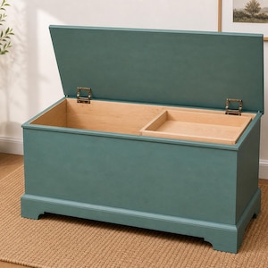 May include: A teal storage chest with an open lid, revealing a natural wood interior and a small tray. The chest has brass hinges and sits on a beige rug. The chest is a rectangular shape.