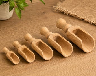 May include: A set of five graduated wooden scoops arranged in a row from smallest to largest. Each scoop features a smooth, light brown natural wood grain, a rounded handle, and a deep, open-top basin for measuring or transferring dry goods.