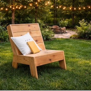 May include: A wooden outdoor chair with a slatted back and a wide seat. A white pillow and a yellow book rest on the chair. The chair is set on a green lawn, with a fire pit and string lights in the background.