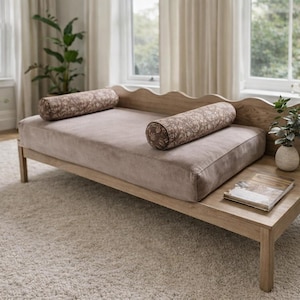 May include: A light wood daybed with a curved back and a built-in side table. The daybed has a light brown cushion and two patterned bolster pillows. A small plant in a vase sits on the side table.