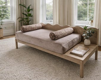 May include: A light wood daybed with a curved back and a built-in side table. The daybed has a light brown cushion and two patterned bolster pillows. A small plant in a vase sits on the side table.