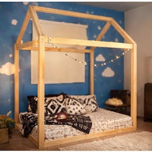May include: A wooden house-shaped bed frame with a mattress and pillows. The bed is decorated with a patterned blanket and pillows. A string of lights is draped across the top of the frame. The background is a blue wall with white cloud and star designs.