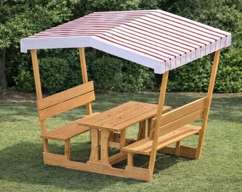 May include: A wooden picnic table set with a striped canopy. The table has attached benches and is made of light-colored wood. The canopy is white with red stripes. The set is outdoors on a grassy lawn.