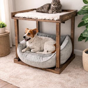 May include: A wooden pet bed with a gray cushion for a dog and a top level with a soft, white surface for a cat. The dog bed is below the cat bed. The frame is made of dark wood.