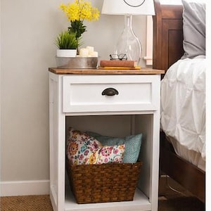 May include: A white nightstand with a wooden top, a drawer, and an open shelf. A woven basket holds pillows with floral and teal patterns. A lamp with a white shade and a black "n" is on top, along with yellow flowers.