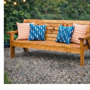 Puede incluir: Un banco de jardín de madera con tres cojines decorativos. Dos cojines tienen un patrón azul y los otros dos tienen un diseño de chevron naranja. El banco está hecho de madera de color marrón claro y está sobre un lecho de piedras pequeñas.