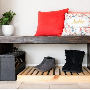 May include: A rustic wooden bench with a dark stain, featuring a lower shelf and a built-in shoe rack. The bench is adorned with a red pillow and a floral pillow with the word "hello". A potted plant and two pairs of shoes complete the scene.
