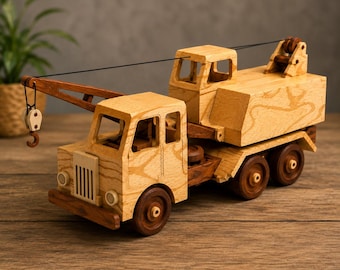 May include: A wooden toy crane truck featuring a light tan cab and a darker brown chassis with six wheels. The vehicle includes a rotating crane arm with a black string and a small hook hanging from the end, plus a detailed grille and headlights.