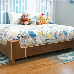 以下が含まれることがあります： カラフルな模様の掛け布団が付いた、ロープロファイルの木製ベッドフレーム。ベッドにはぬいぐるみがあしらわれ、木製の床の上に置かれています。部屋には青と白のストライプの壁があります。