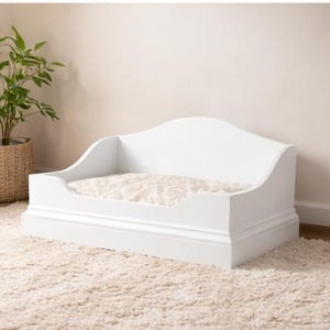 May include: A white wooden pet bed with a decorative headboard and base. The bed features a patterned cushion and is designed for small pets. The bed is placed on a cream-colored rug, with a potted plant in the background.