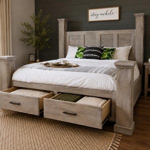 May include: A light gray wooden bed frame with two open drawers. The bed has a headboard with a sign that says "stay awhile". The bed is made with white bedding and decorative pillows. A brown rug is in front of the bed.