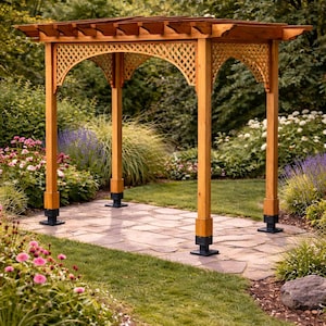 May include: A wooden garden arbor with a decorative latticework design. The arbor stands on a stone pathway, surrounded by a garden with green grass and colorful flowers. The structure has four supporting posts and a slatted roof, creating a shaded area.