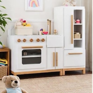 May include: A white wooden play kitchen set with a refrigerator, oven, and sink. The set has wooden accents and gold-colored hardware. A framed rainbow picture hangs on the wall. A teddy bear sits on the floor.