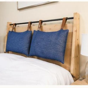 May include: A wooden headboard with two blue cushions suspended from a black bar by brown leather straps. The headboard is made of light-colored wood and is positioned above a white bed. A lamp is visible on the right.