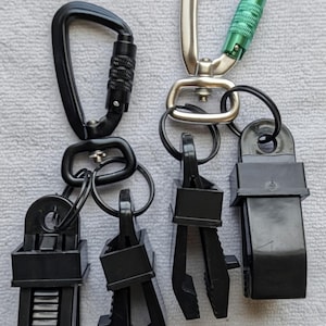 May include: Two carabiner clips with black and silver metal hardware. One black carabiner has a screw lock, and the other has a green accent. Each clip is attached to a black plastic clamp.