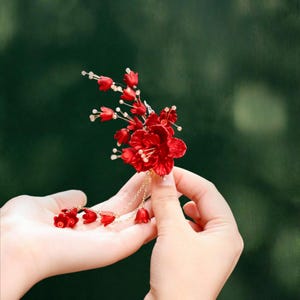 May include: A vibrant red floral hair accessory with delicate gold chain accents. The handcrafted piece features multiple red flower blossoms and smaller buds, creating a layered and textured design. The accessory is held against a blurred green background.