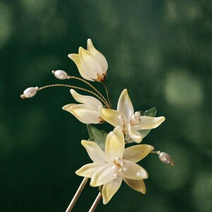 May include: A floral hair accessory featuring delicate, pale yellow flower petals with pearl accents. The flowers are attached to a gold-toned metal hair pin, creating an elegant and natural look.