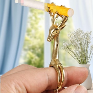 Könnte beinhalten: Ein goldfarbener zigarettenhalterförmiger Ring in Hirschform. Der Ring ist so konzipiert, dass er eine Zigarette hält, wobei das Geweih des Hirsches die Zigarette umschließt. Der Ring wird am Finger getragen, und der Kopf des Hirsches ist aufrecht. Der Hintergrund ist unscharf.