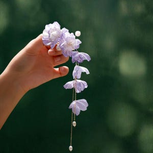May include: A hair accessory featuring multiple light purple fabric flowers arranged in a cascading design. The piece includes thin gold-toned metal chains hanging downwards, accented with small white beads and pearls at the ends.