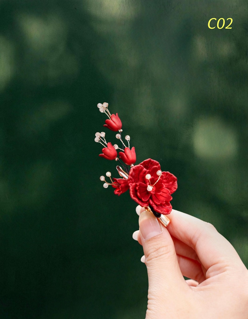 Op de afbeelding: Een rode bloemenbroche met kleine witte accenten en goudkleurige metalen details. De broche heeft een cluster van rode bloemen en knoppen, met de tekst "C02" in de rechterbovenhoek.