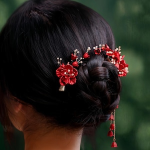 Op de afbeelding: Close-up van donker haar in een knot, versierd met felrode bloemen haaraccessoires. De accessoires omvatten kleine rode bloemen, witte parelachtige kralen en bungelende rode klokvormige bedels, wat een elegante en feestelijke uitstraling cre&euml;ert.