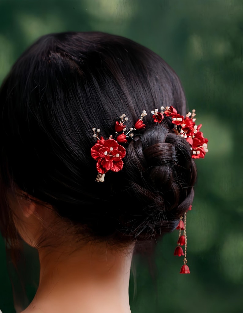Op de afbeelding: Close-up van donker haar in een knot, versierd met felrode bloemen haaraccessoires. De accessoires omvatten kleine rode bloemen, witte parelachtige kralen en bungelende rode klokvormige bedels, wat een elegante en feestelijke uitstraling cre&euml;ert.