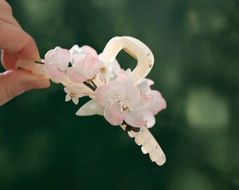 May include: A cream-coloured hair claw clip adorned with delicate, multi-layered pink and white flower blossoms. The floral embellishments feature soft gradient petals and small white centres, securely attached to the top of the functional hair accessory.