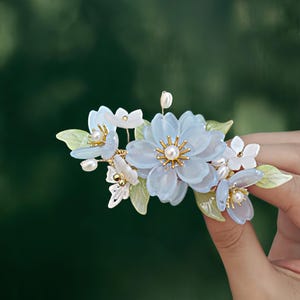May include: A floral hair clip featuring light blue and white flowers with gold accents and faux pearls. The clip has delicate green leaves and is designed to be worn in the hair. The flowers have a translucent appearance.